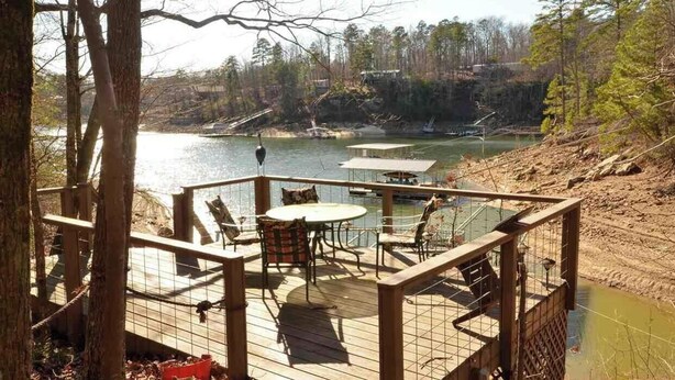 Une terrasse en bois avec une table et des chaises, donnant sur un lac, entourée d’arbres et bordée d’une rive rocheuse.