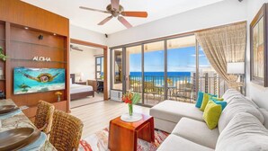 Een moderne woonkamer met een grote televisie, een plafondventilator, een balkon met uitzicht op de oceaan en een eethoek met rieten stoelen.