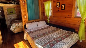 Ein Schlafzimmer einer Holzhütte mit einem großen Bett, einem kleinen angrenzenden Schlafbereich, einem Deckenventilator und einem Fenster mit Vorhängen.