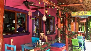 A colorful outdoor seating area with tables and chairs, a sign for 'Little Day Cafe & Bakery', and a thatched roof structure. - Oceanfront Penthouse -4300 Sq Ft, 2 Level -Mi Casa Del Mar (Akumal)