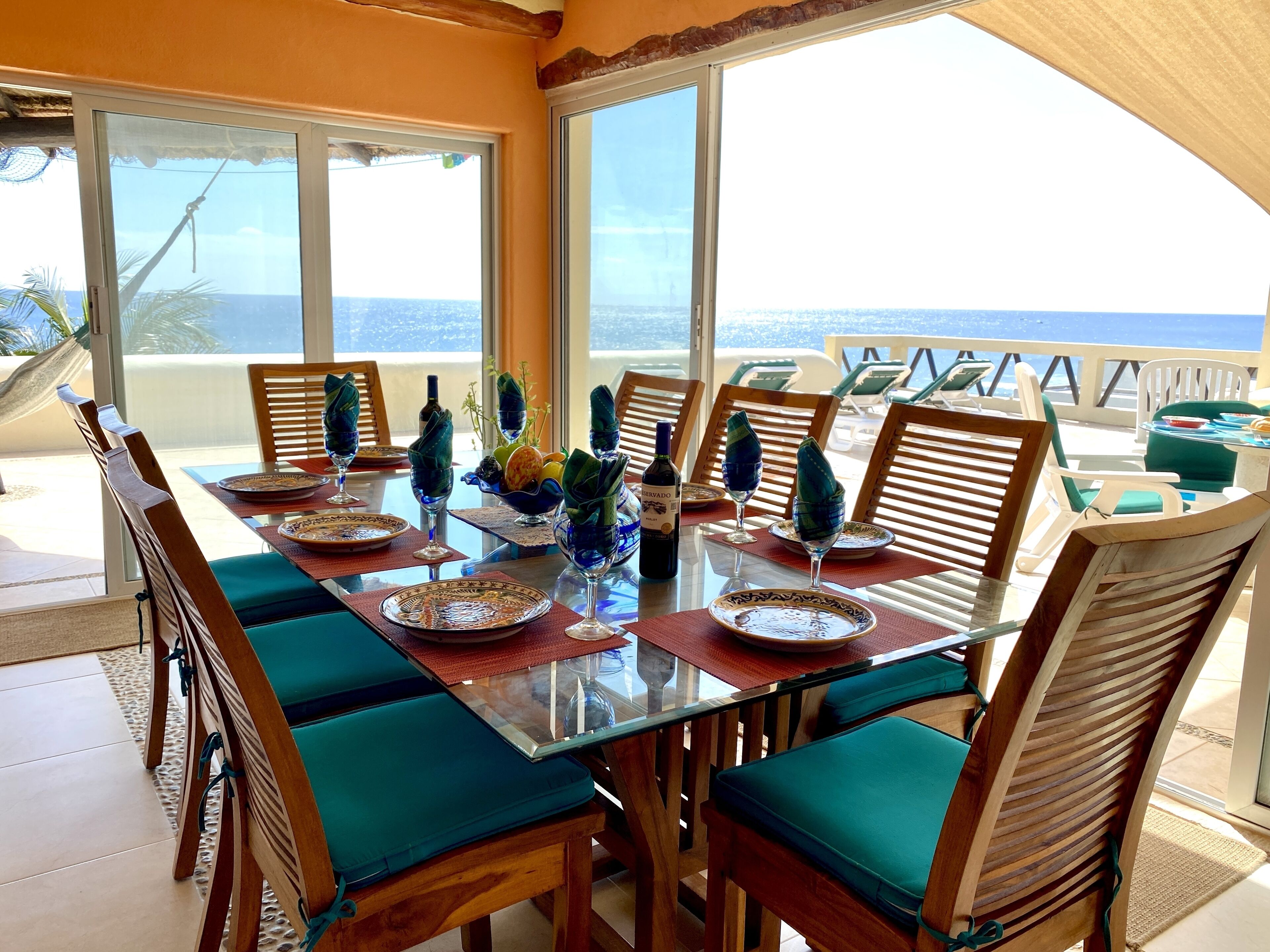 Un tavolo da pranzo apparecchiato con piatti, bicchieri e una bottiglia, circondato da sedie di legno con cuscini verdi, su un balcone con vista sull'oceano.