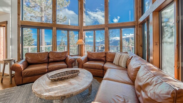 A spacious living room with large windows, leather sofas, and a rustic coffee table. - Tahoe Point of View by Tahoe Getaways - Hot Tub + Sauna + Lake Views + Private Beach (Tahoe City)