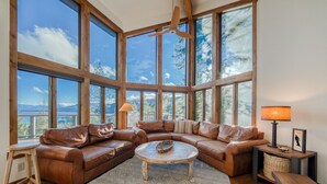 A spacious living room with large windows, a leather sectional sofa, a round coffee table, and a wooden ceiling fan. - Tahoe Point of View by Tahoe Getaways - Hot Tub + Sauna + Lake Views + Private Beach (Tahoe City)
