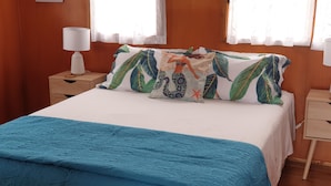 Een net opgemaakt bed met een blauw sprei, een wit kussen met een tropisch bladpatroon, en een passend kussen op een houten nachtkastje.