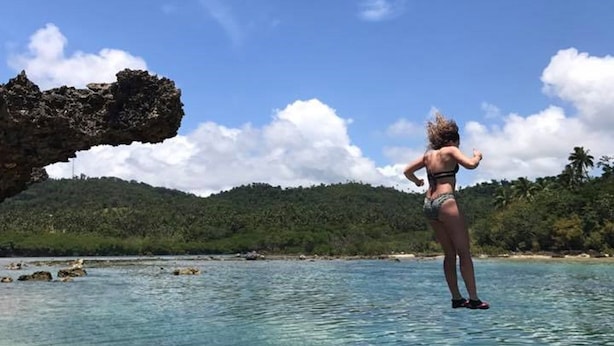 Een vrouw in een bikini staat op een rotsachtige kust met helderblauw water en een grote rotsformatie op de achtergrond.