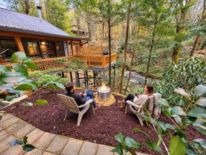 A wooden deck with two chairs and a fire pit, surrounded by a forested area.