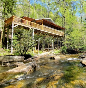 A wooden cabin with a balcony overlooking a clear stream, surrounded by trees.