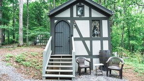 A small wooden cabin with a dark door, white trim, and a window. There are two chairs and a table outside. - #8 Helga - Tiny Home Cottage (Sevierville)