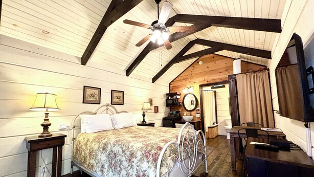 A bedroom with a wooden ceiling, a bed with a floral bedspread, a nightstand with a lamp, a dresser, and a mirror. - #8 Helga - Tiny Home Cottage (Sevierville)
