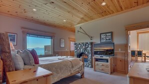 A cozy bedroom with a bed, a stone fireplace, a television, and a view of the outdoors.