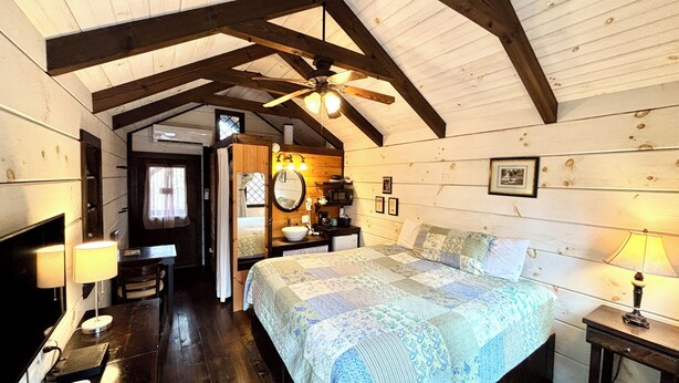 Une chambre confortable dans un chalet, avec un plafond en bois, un lit recouvert d’une courtepointe, une table de chevet, une lampe et un ventilateur de plafond.