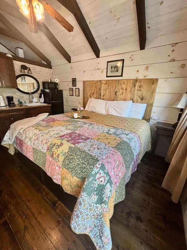 A bedroom with a wooden ceiling, a bed with a floral quilt, a nightstand, a dresser, a mirror, and a framed picture on the wall. - #4 Stella - Tiny Home Cottage (Sevierville)