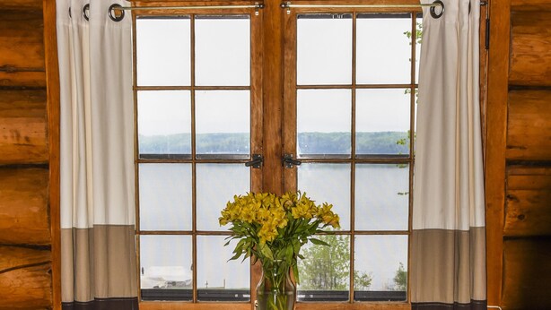 Et interiør af en træhytte med et rundt bord og to stole, en vase med gule blomster samt udsigt til en sø gennem et vindue med gardiner.