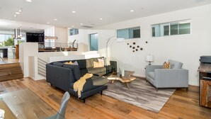 Un vaste espace de vie comprenant un canapé gris foncé, un fauteuil gris clair et une table basse en verre. Une table à manger en bois est placée au premier plan.