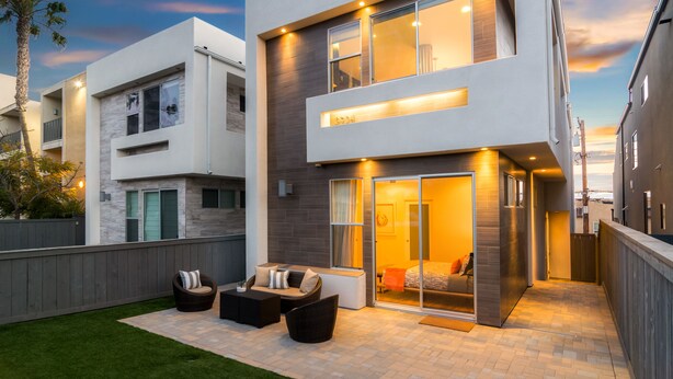 Une maison moderne à deux étages dotée d’un patio aménagé avec du mobilier d’extérieur et une porte-fenêtre coulissante donnant accès à une chambre.