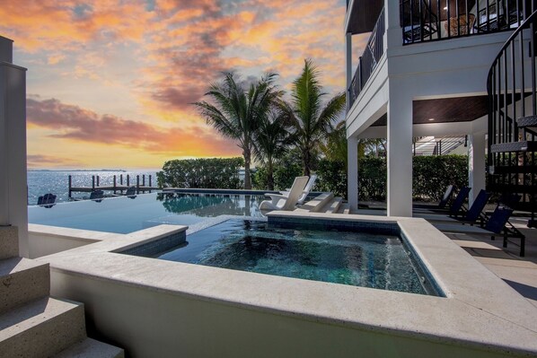 A swimming pool with a view of the ocean and palm trees, surrounded by lounge chairs and a multi-level building.
