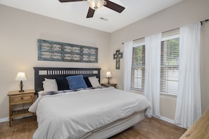 A bedroom with a bed, two bedside tables with lamps, a ceiling fan, a window with blinds, and a wall decoration.