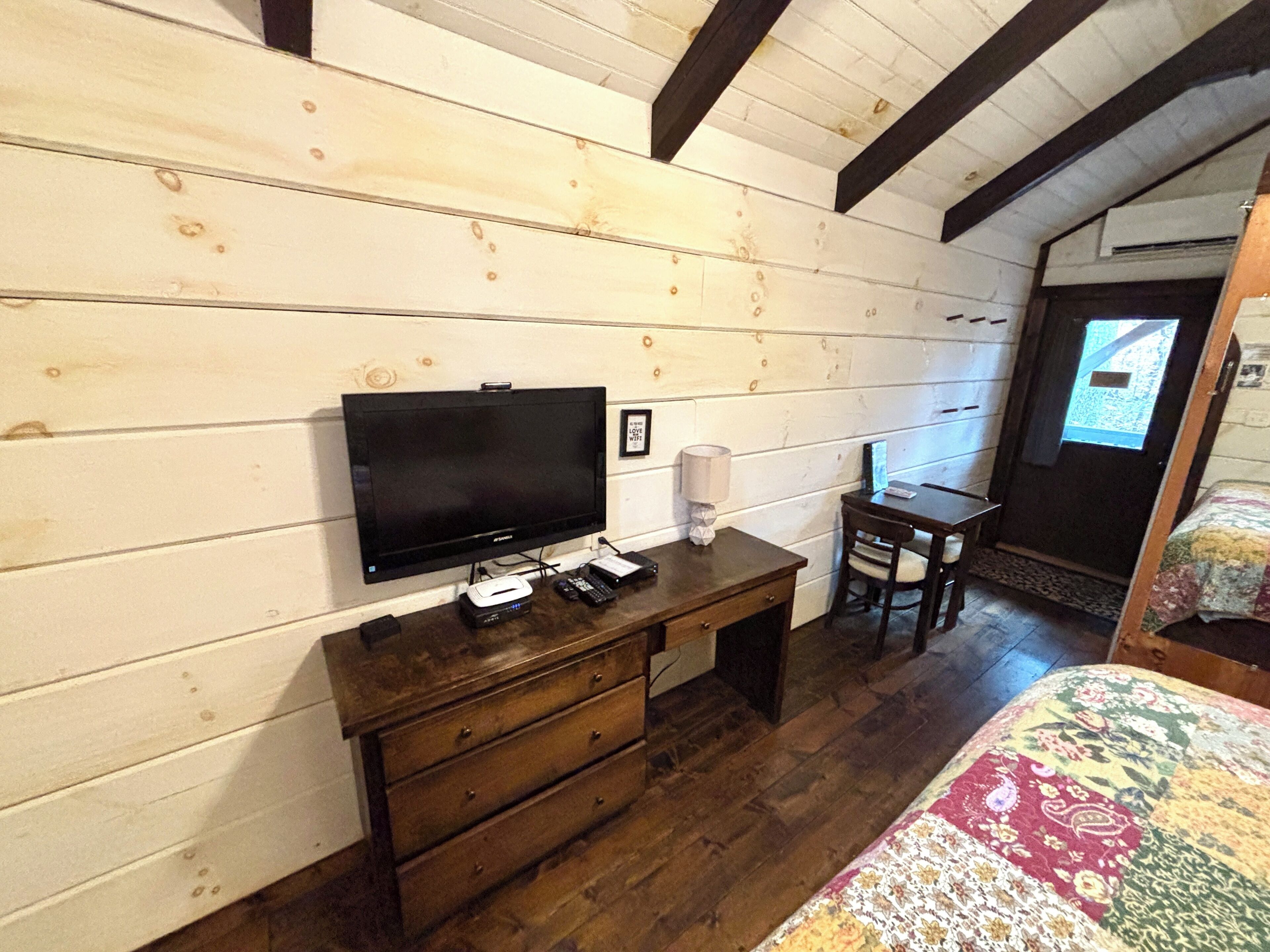 Ein Zimmer mit Holzwänden, einem Fernseher auf einer Holzkommode, einem Schreibtisch mit Stuhl und einem Bett mit gemustertem Überwurf.