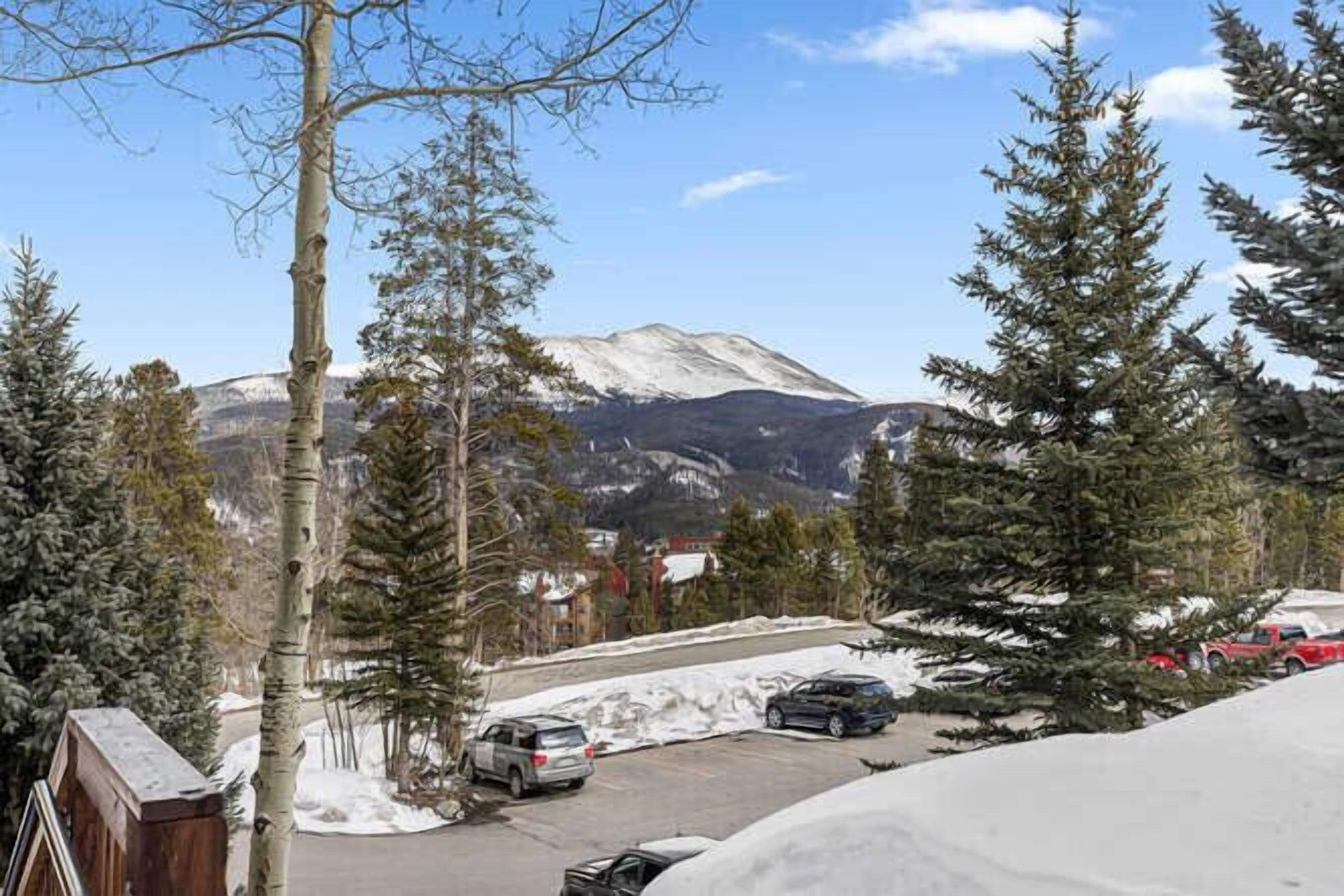 A parking area with snow-covered ground, surrounded by tall evergreen trees and a mountain in the background.