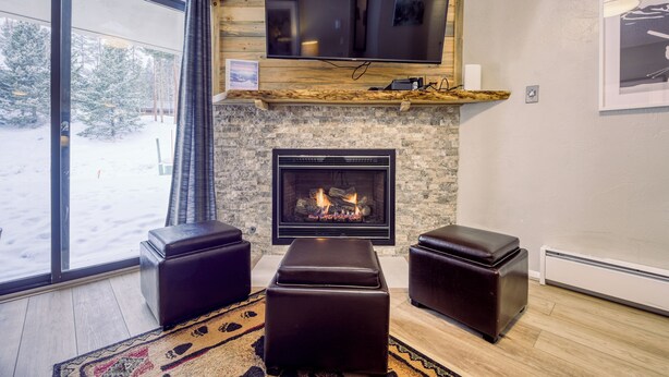 Una sala de estar con chimenea, un televisor montado en la pared y un amplio ventanal con vista a árboles.