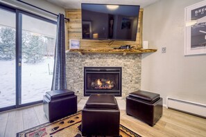 A living room with a fireplace, a television mounted on the wall, and a large window with a view of trees. - Ski in/ Ski out 1st Floor Condo in Breckenridge (Breckenridge)