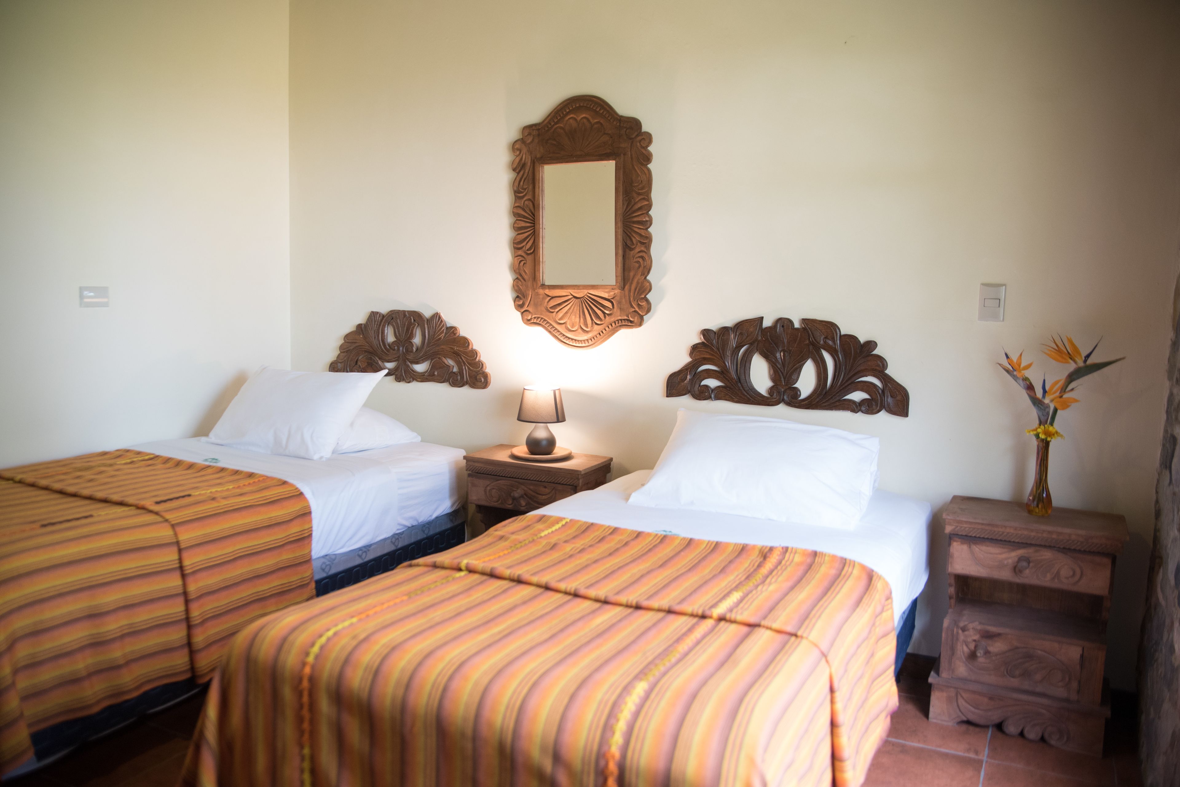 A hotel room with two beds, a mirror, a nightstand with a lamp, and a small wooden cabinet.