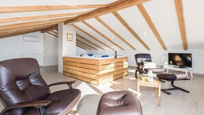 Ein geräumiger Loftsraum mit freiliegenden Holzbalken, einer zentralen Holztischinsel, Ledersesseln, einem Fernseher und einem kleinen Tisch mit einer Laterne.