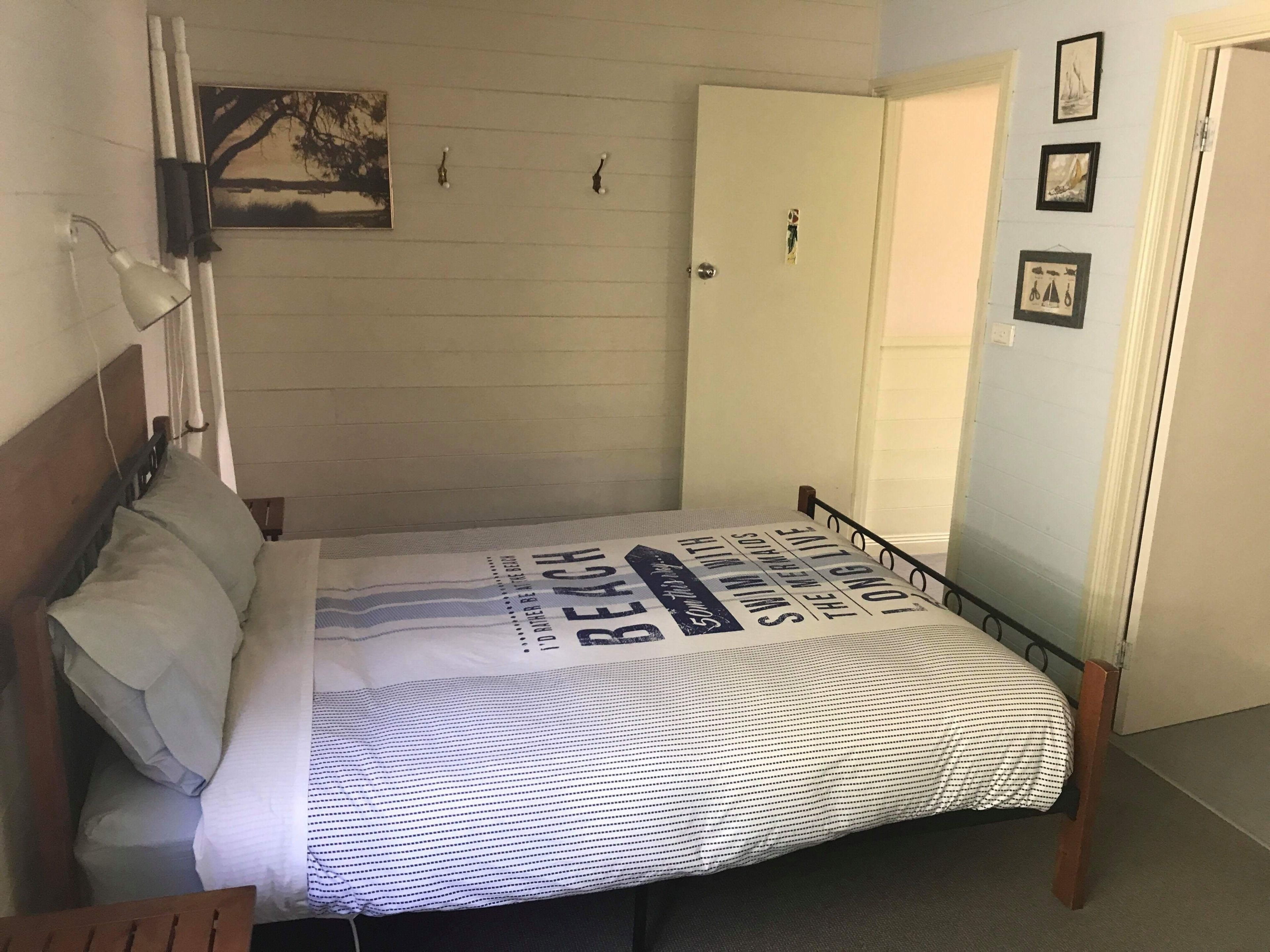 Une chambre à coucher avec un lit, une table de chevet, une lampe fixée au mur, une porte et un tableau encadré.