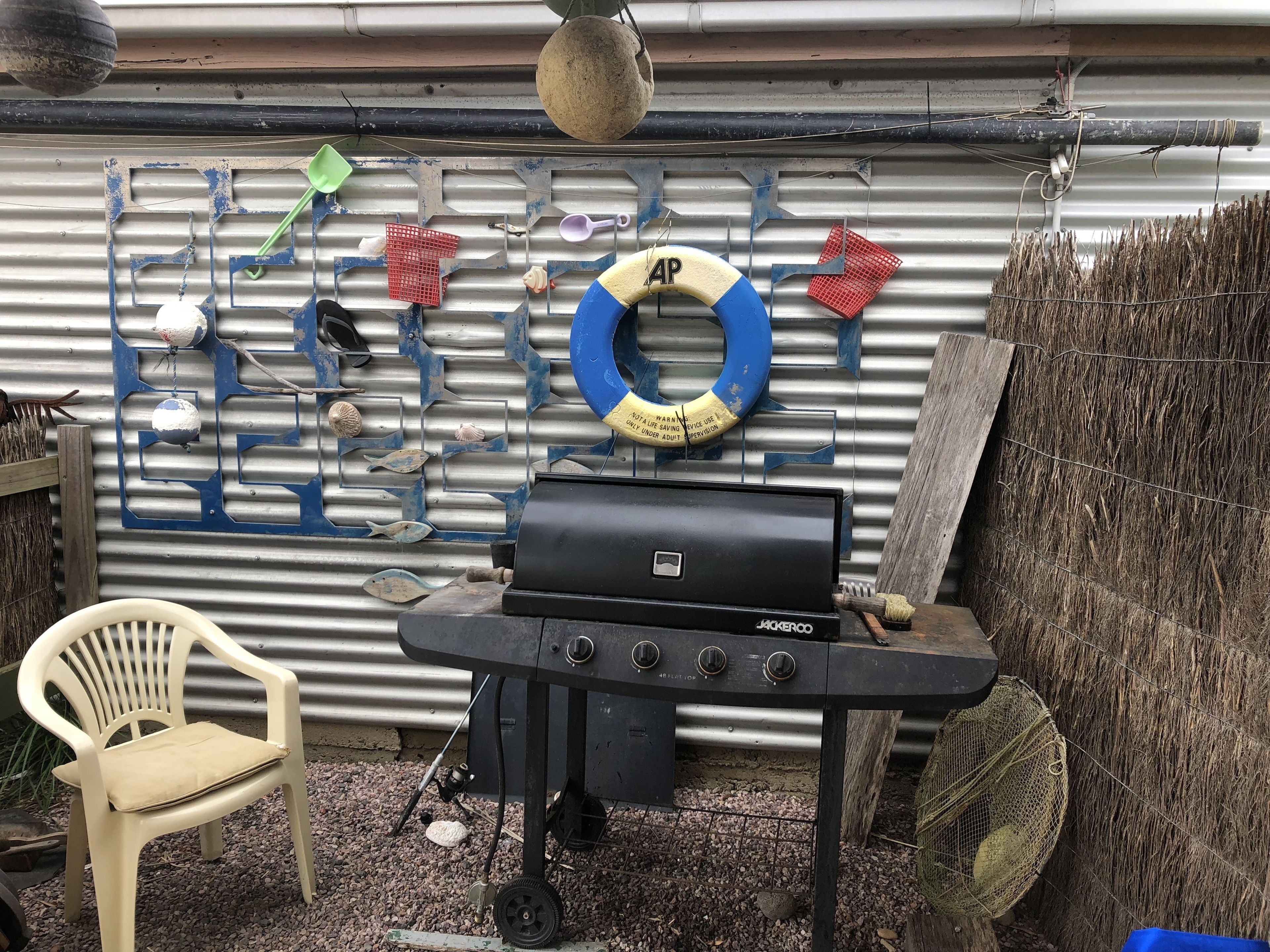 Una parrilla para barbacoa en un entorno exterior, con un muro de metal corrugado decorado con varios objetos como un flotador, conchas marinas y una pala verde.