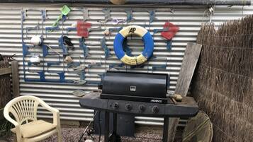 Una parrilla para barbacoa en un entorno exterior, con un muro de metal corrugado decorado con varios objetos como un flotador, conchas marinas y una pala verde.