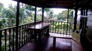 Una terraza de madera con una mesa y bancos, rodeada por una barandilla, con vistas a un jardín exuberante.