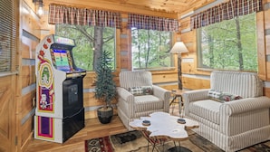 Un acogedor interior de cabaña con una máquina de pinball, dos sillones a rayas, una mesita y un árbol de Navidad.