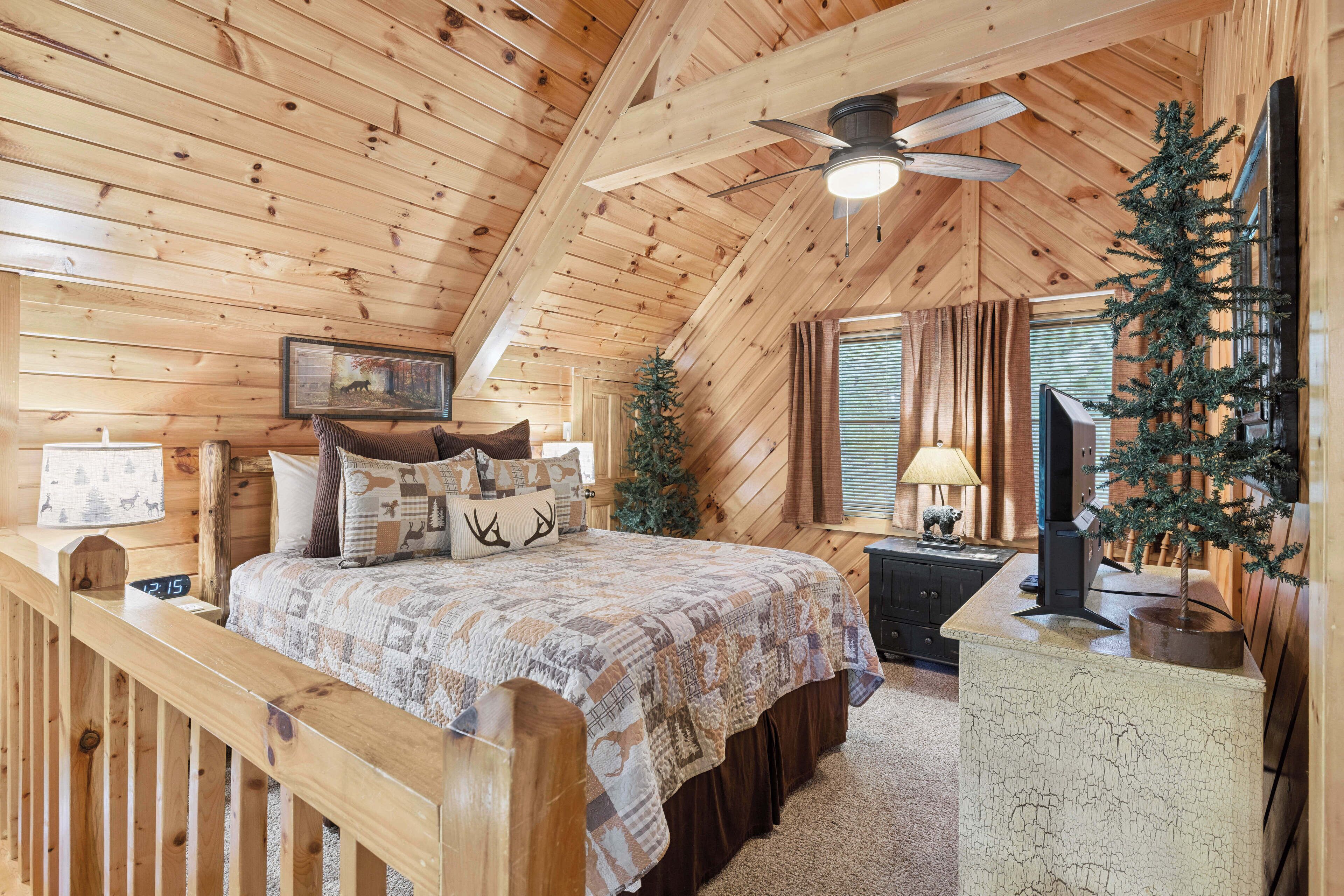Un dormitorio de cabaña de madera con cama, un pequeño árbol de Navidad, un ventilador de techo y una ventana con cortinas.