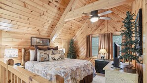 Un dormitorio de cabaña de madera con cama, un pequeño árbol de Navidad, un ventilador de techo y una ventana con cortinas.
