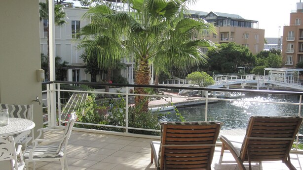 Un balcon avec deux fauteuils de jardin en bois, une petite table sur laquelle se trouve un bocal en verre, et une vue sur un canal, avec des bâtiments et un pont en arrière-plan.
