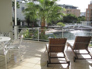 A balcony with two wooden lounge chairs, a small table with a glass jar, and a view of a canal with buildings and a bridge in the background. - Luxurious, Spacious & Secure V&A Waterfront Apt (Cape Town)