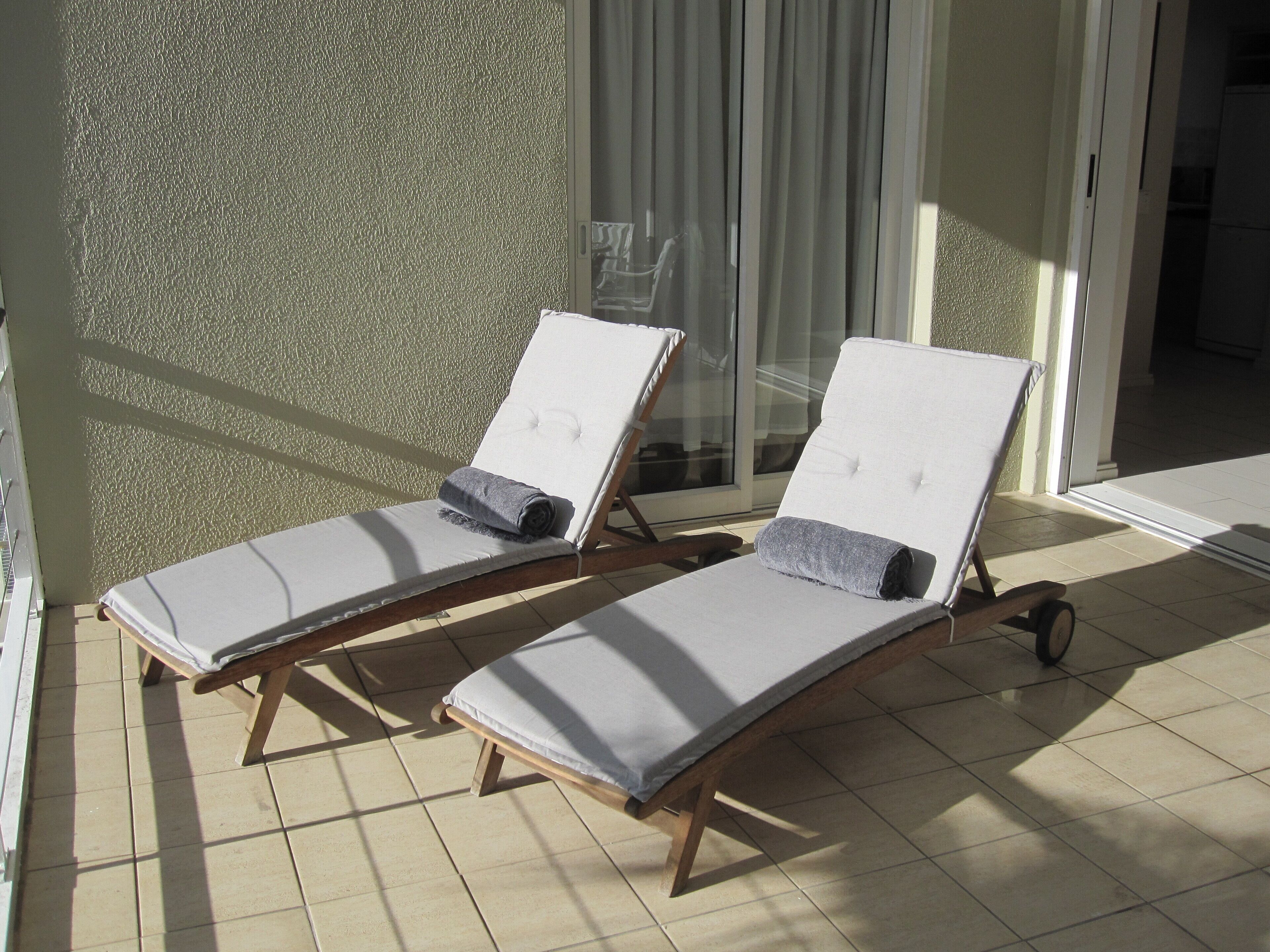 Two sun loungers with cushions, a textured wall, and a glass door leading to a shaded area.