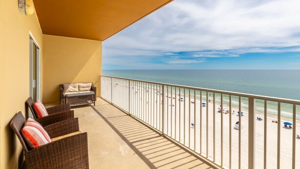 Un balcone con mobili in vimini e vista su una spiaggia con persone e ombrelloni.