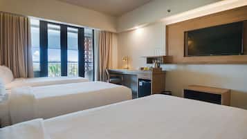 A hotel room with two beds, a desk, a small kitchenette, and a large window with curtains.
