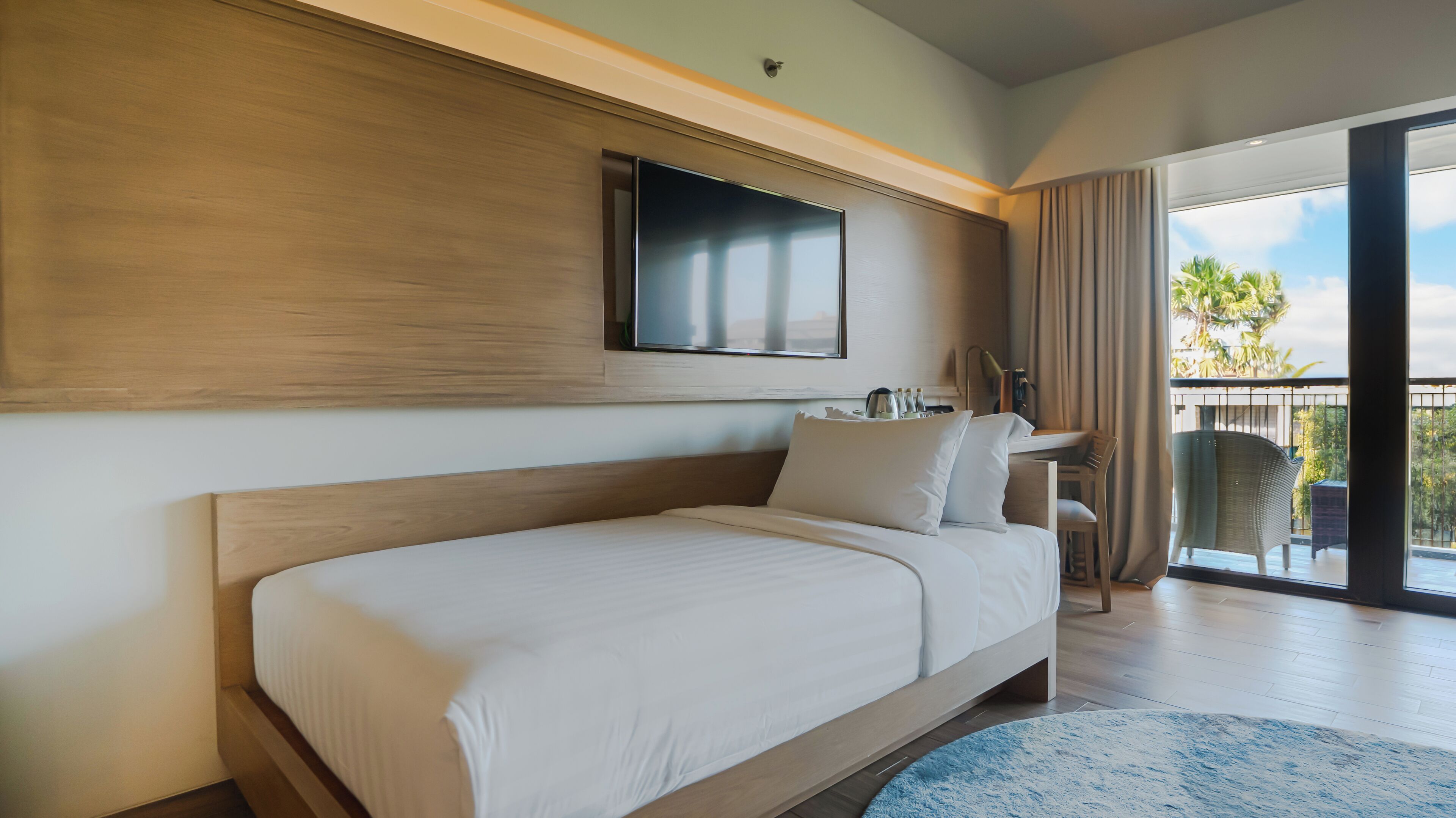 A modern hotel room with a large bed, a flat-screen TV mounted on the wall, a desk with a chair, and a balcony with a view of palm trees.