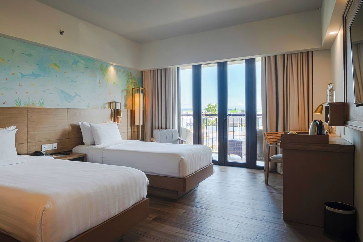 A hotel room with two beds, a large mural, a balcony with curtains, and a view of the outdoors.