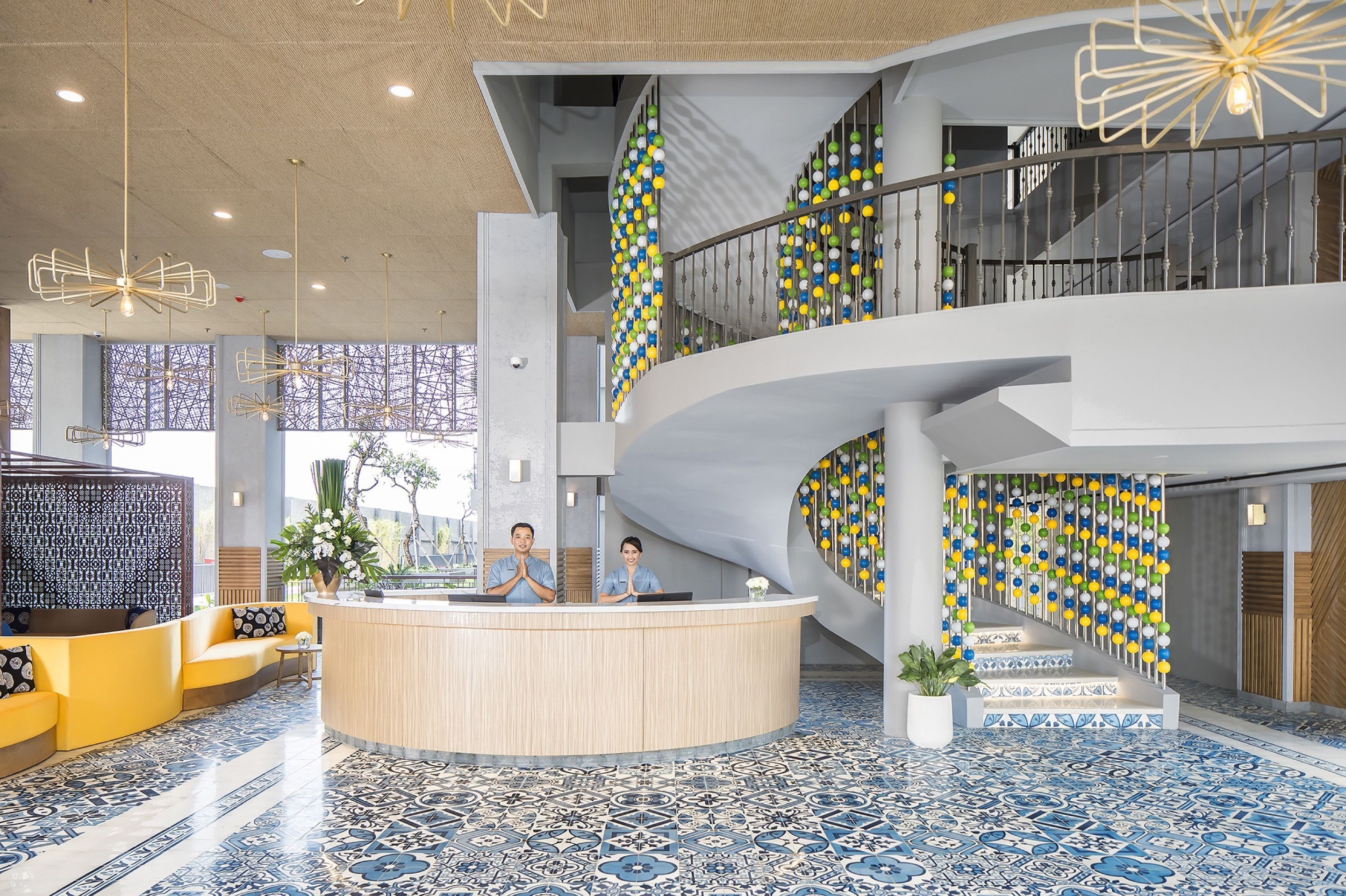 A modern hotel lobby with a central reception desk, seating areas, and a staircase with colorful decorations.