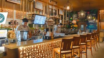 Zona de bar con mostrador, taburetes y una pantalla de televisión mostrando un programa. Hay estantes con botellas y vasos, y una pared decorativa con motivos de café.