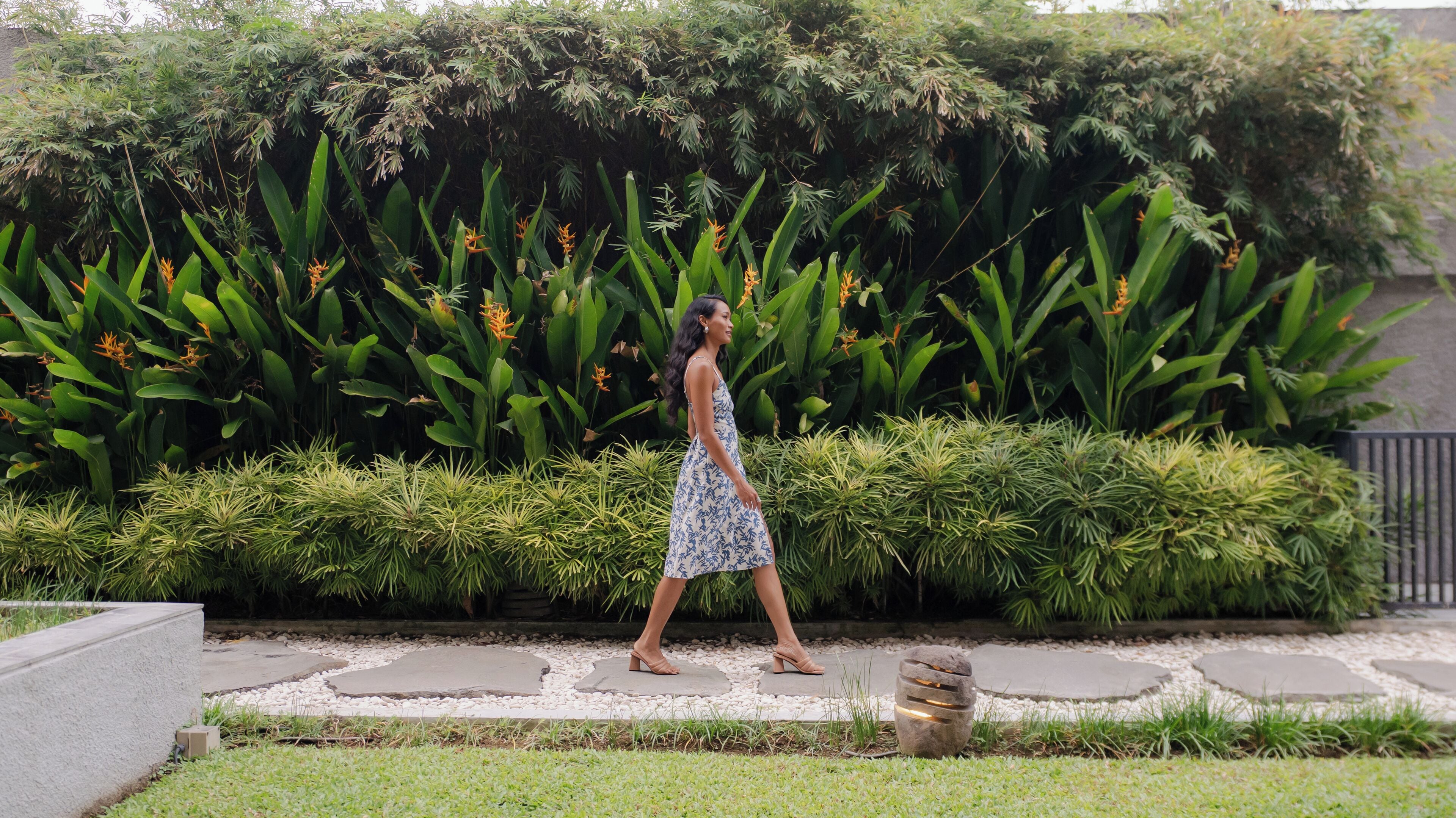 A woman walking on a stone pathway surrounded by lush greenery and tropical plants.