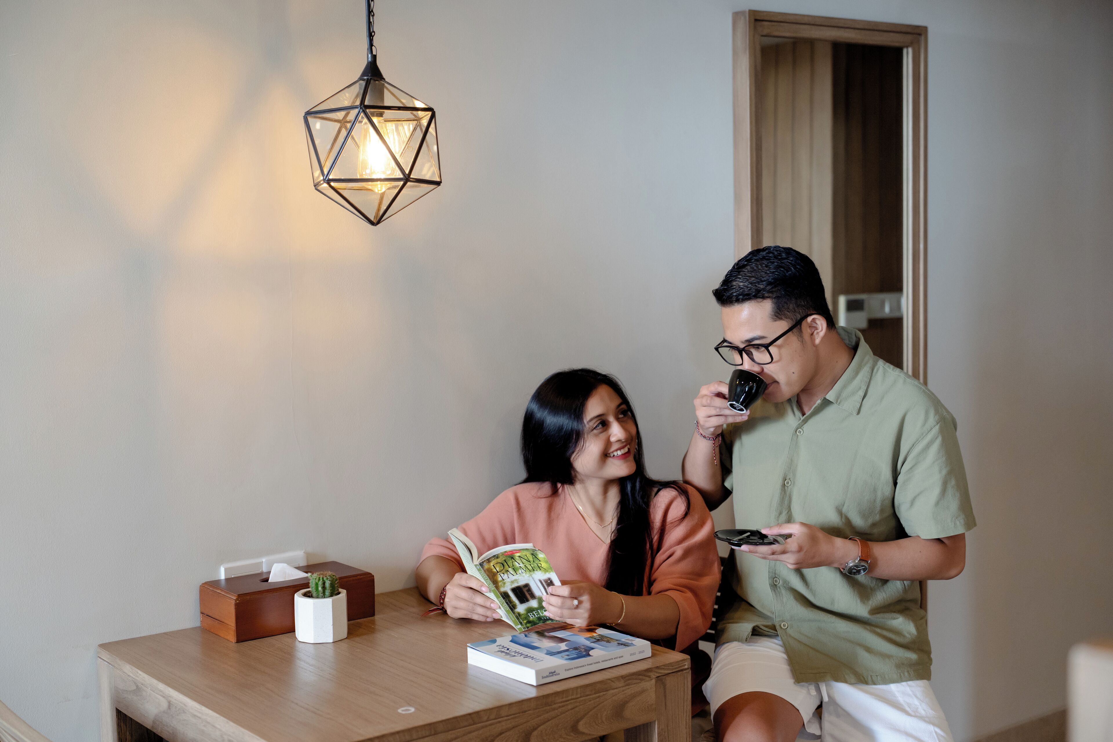 Un couple est assis à une table en bois, entouré de livres et d’une petite plante, dans une pièce éclairée par une suspension aux formes géométriques et dotée d’une porte en bois.