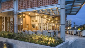 Een buiten eethoek met rieten stoelen en tafels, een houten omheining en hangende rieten lampen.