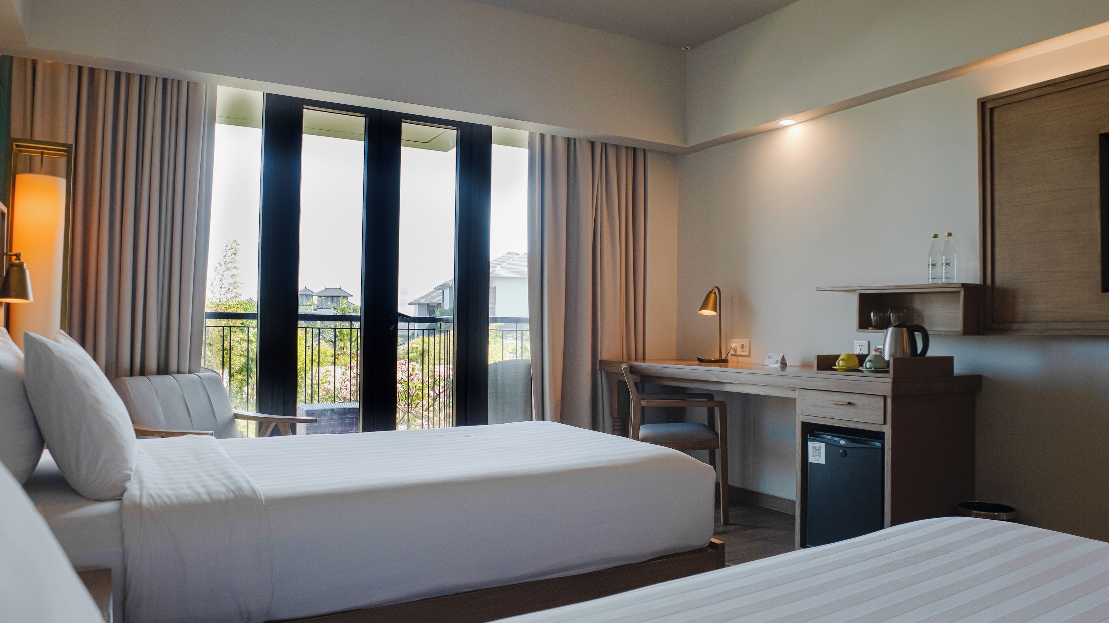 A hotel room with two beds, a desk, a chair, a small fridge, and a balcony with curtains.