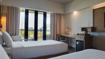 A hotel room with two beds, a desk, a chair, a small fridge, and a balcony with curtains.