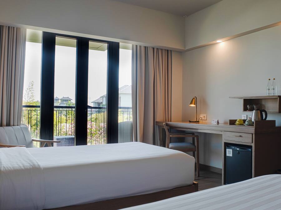 A hotel room with two beds, a desk, a chair, a small fridge, and a balcony with curtains.