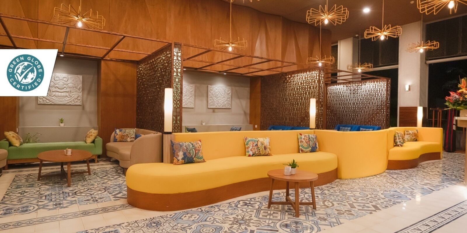 A modern lounge area with a curved yellow sofa, a green sofa, and a small round table. There are decorative panels and a patterned floor.